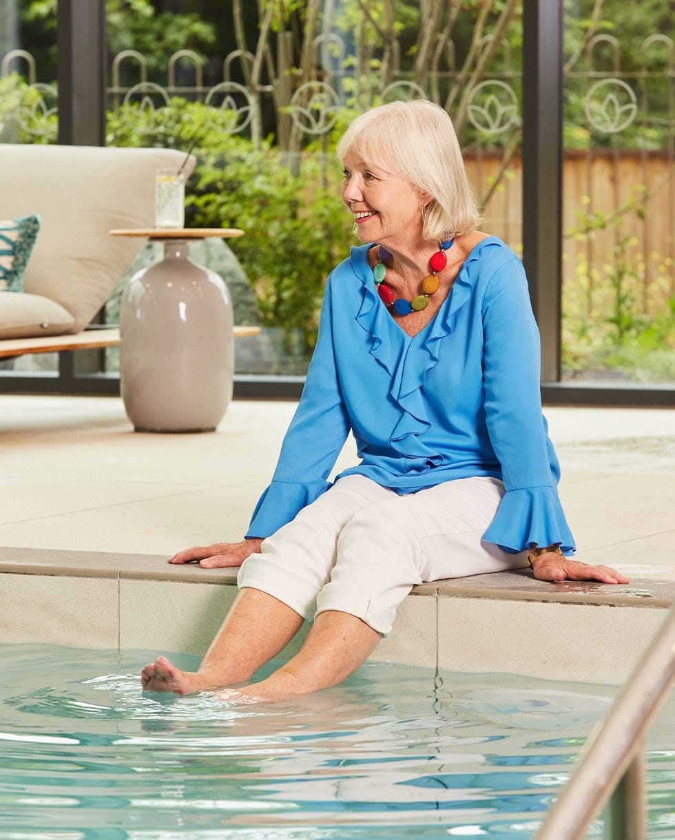 an older woman sits by a swimming pool dipping her feet in the water
