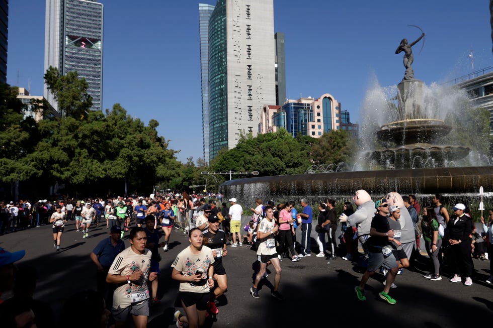 a group of runners pass a fountain with the mexico city skyline in the background