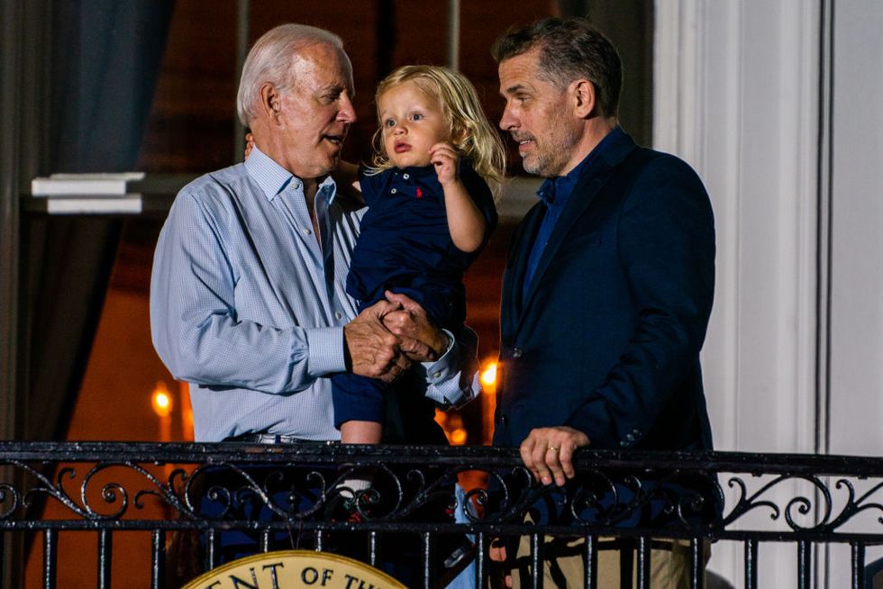 joe biden, wearing a blue dress shirt, holds his young grandson beau while standing next to hunter biden, who wears a dark blue suit jacket, as they stand in front of a railing