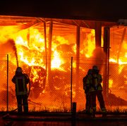 warehouse with hay burns as firefighters stand outside near a fence