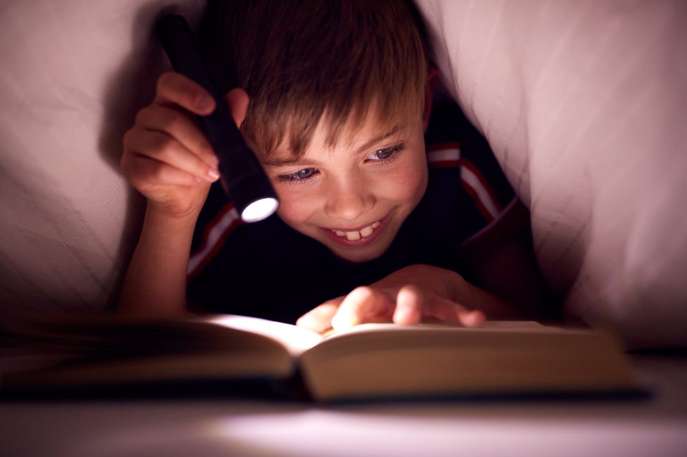 boy at home in bed under blankets reading book whilst holding a flashlight