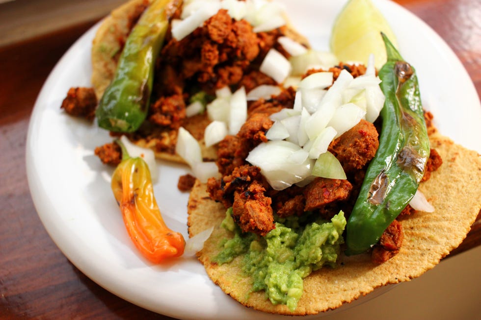 soy chorizo chickpea flour tortillas with blistered shishito peppers, topped with chopped white onions and guacamole on a white plate