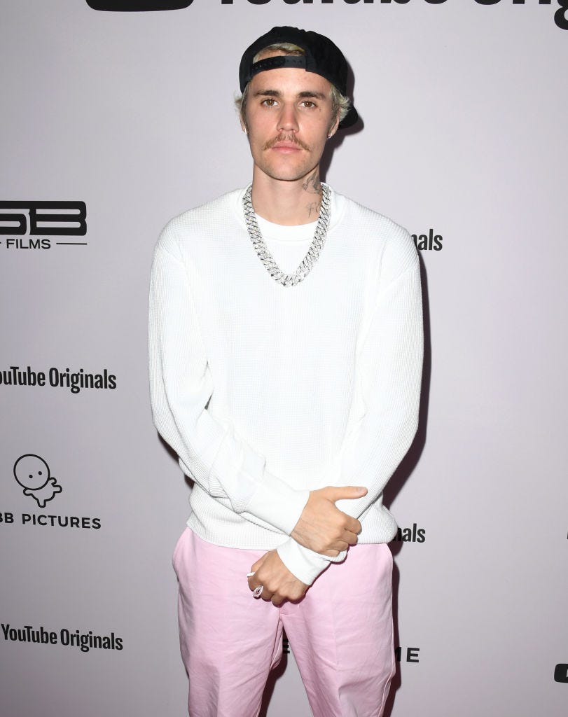 Justin Bieber stands on the red carpet wearing a white sweatshirt, silver chain necklace, pink pants and a backwards black baseball cap with a mustache