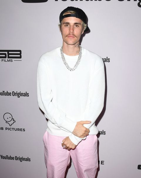 justin bieber stands on the red carpet, wearing a white sweatshirt, silver chain necklace, pink pants, and a black backwards baseball cap he has a mustache