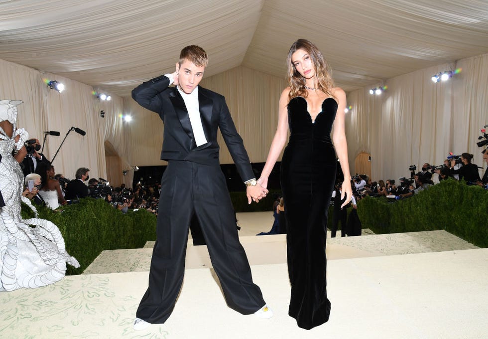 justin bieber and hailey bieber hold hands while standing at the top of a flight of stairs, they look at the camera and wear black formal attire