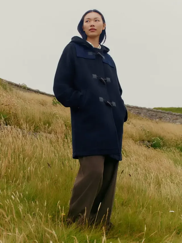 person standing in a grassy field wearing a dark coat and loose pants