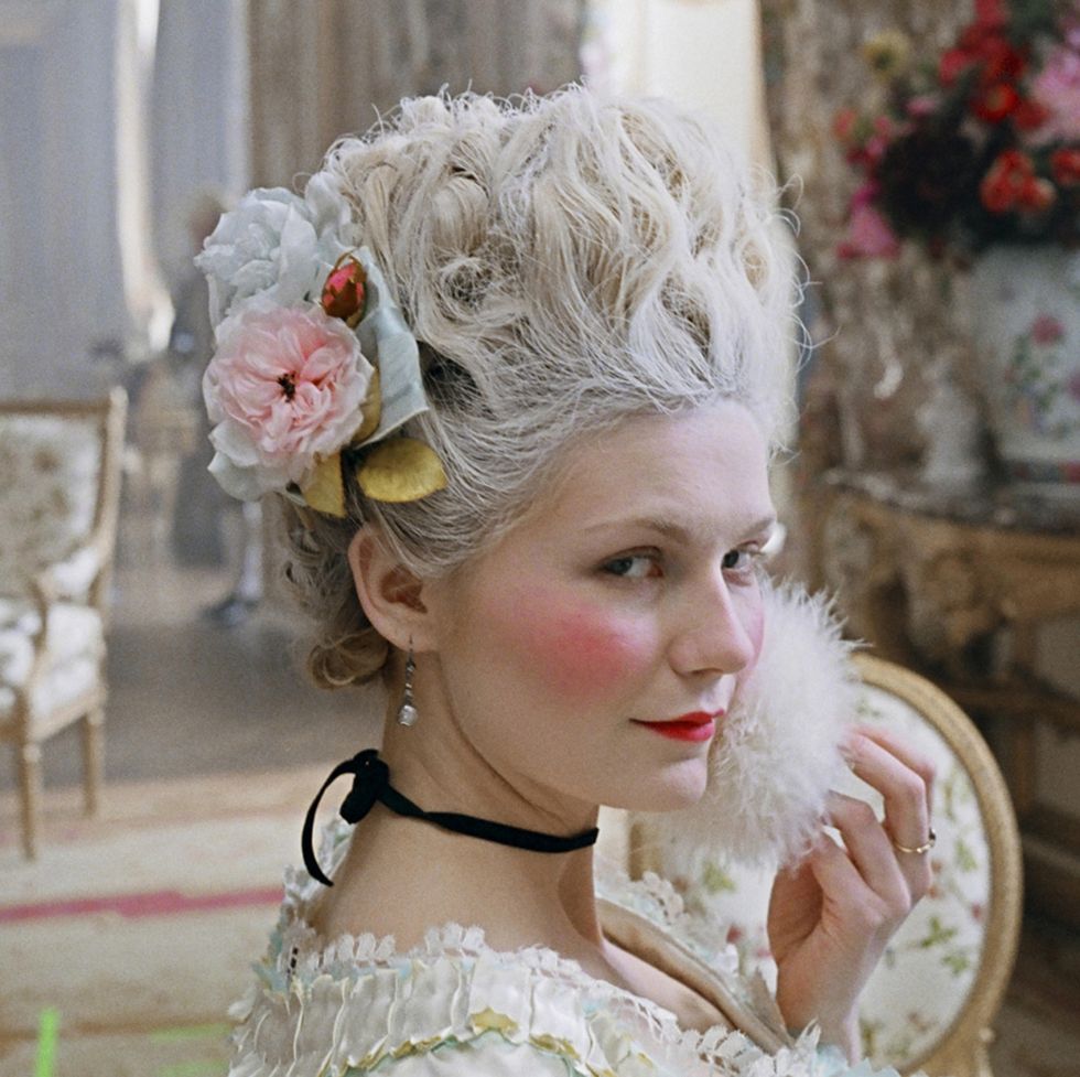 kirsten dunst as marie antoinette sits in a lavish room with a large pink floral bouquet and furniture behind her, she wears a lacy dress, black necklace and flowers in her hair, she holds a white puff to her face as she eyes the camera