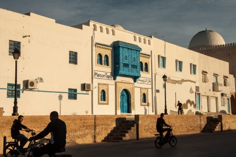 Fietsers rijden langs een tapijten en souvenirwinkel met blauwe deuren in de stad Kairouan Tunesi De beroemdste moskeen in deze heilige stad de Grote Moskee en de Moskee van de Drie Deuren dateren uit 670 en liggen maar vijf minuten fietsen van elkaar