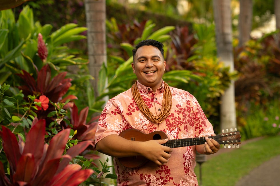 kamahiwa kawaa wearing hawaiian shirt and beaded necklace, holding ukelele, standing in a lush tropical landscape