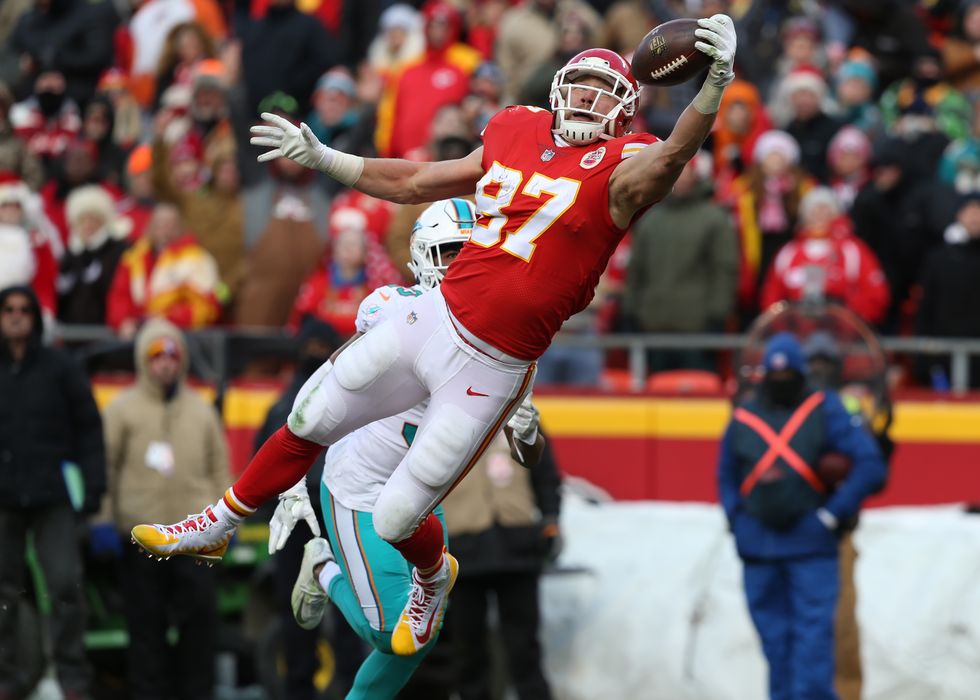 travis kelce, wearing a kansas city chiefs football uniform, jumping in the air and trying to catch a football with one hand