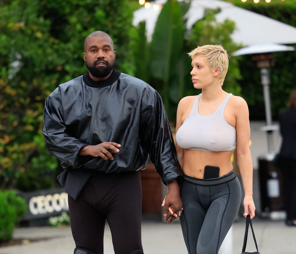 kanye west and bianca censori walk hand in hand outside, he wears all black, she wears a white sports bra and gray leggings