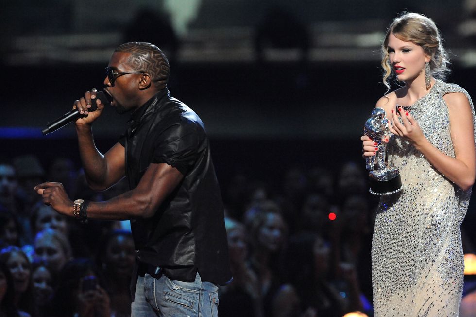 kayne west speaks into a handheld microphone on a stage as taylor swift stands feet away holding a silver trophy, he wears a black shirt with jeans and sunglasses, she wears a bejeweled one strap dress