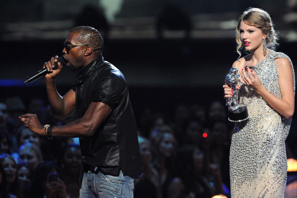 kayne west speaks into a handheld microphone on a stage as taylor swift stands feet away holding a silver trophy, he wears a black shirt with jeans and sunglasses, she wears a bejeweled one strap dress