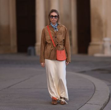 vrouw in bruine jas oranje tas en lange witte rok