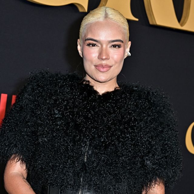 karol g smiles at the camera, she wears a fluffy black outfit and stands in front of a black background with gold and red writing on it