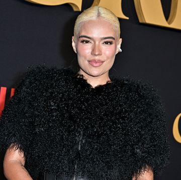 karol g smiles at the camera, she wears a fluffy black outfit and stands in front of a black background with gold and red writing on it