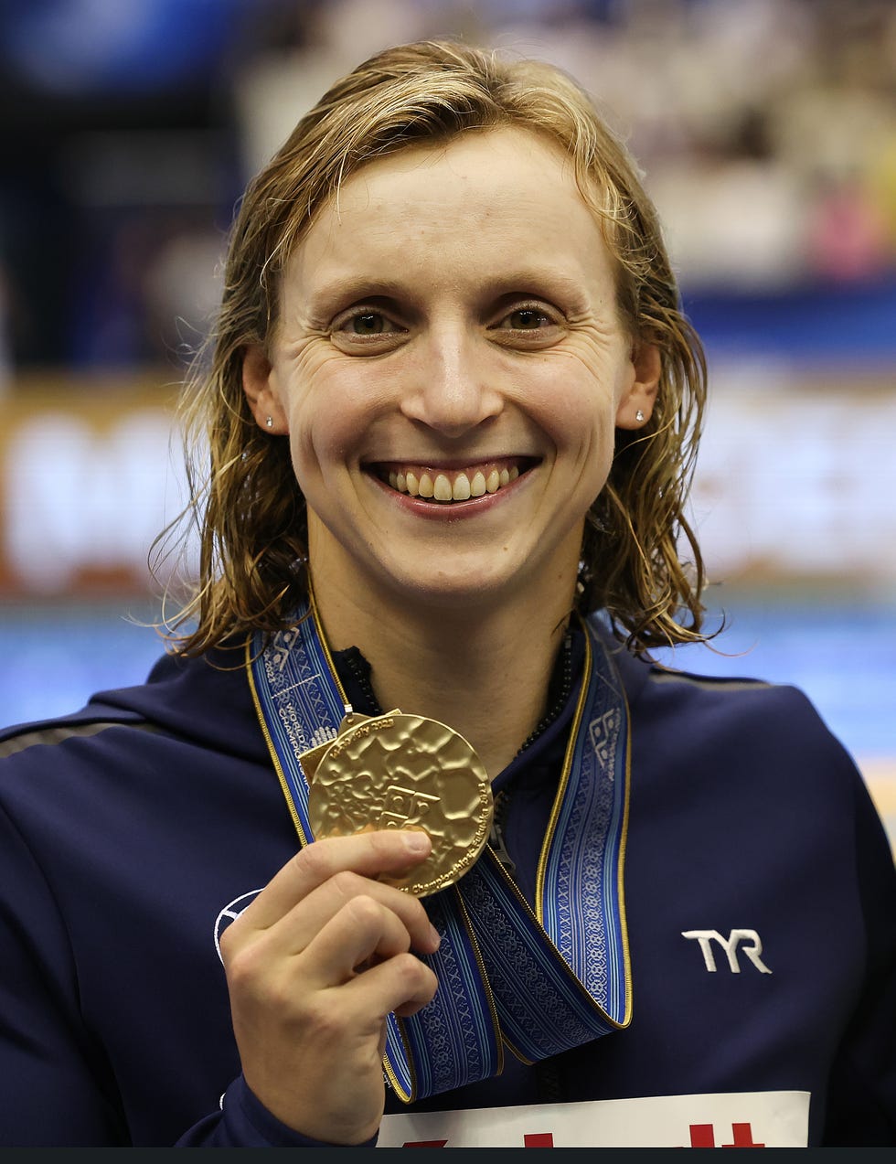 katie ledecky smiling and holding out a gold mdal with her right hand