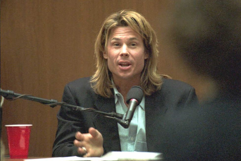 kato kaelin speaks while sitting behind a microphone, he wears a suit jacket and green collared shirt