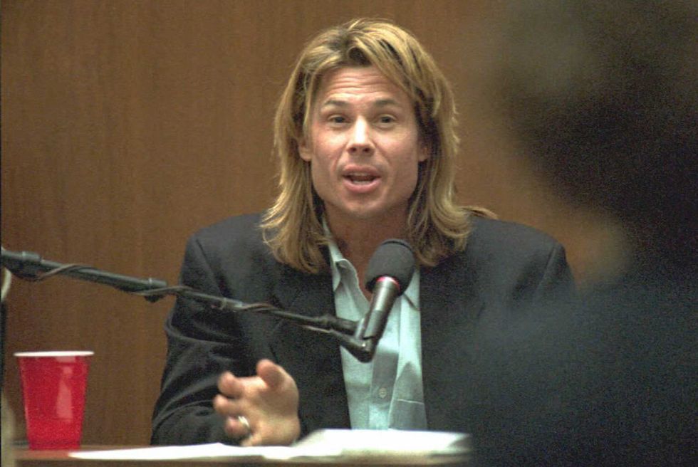kato kaelin speaks while sitting behind a microphone, he wears a suit jacket and green collared shirt