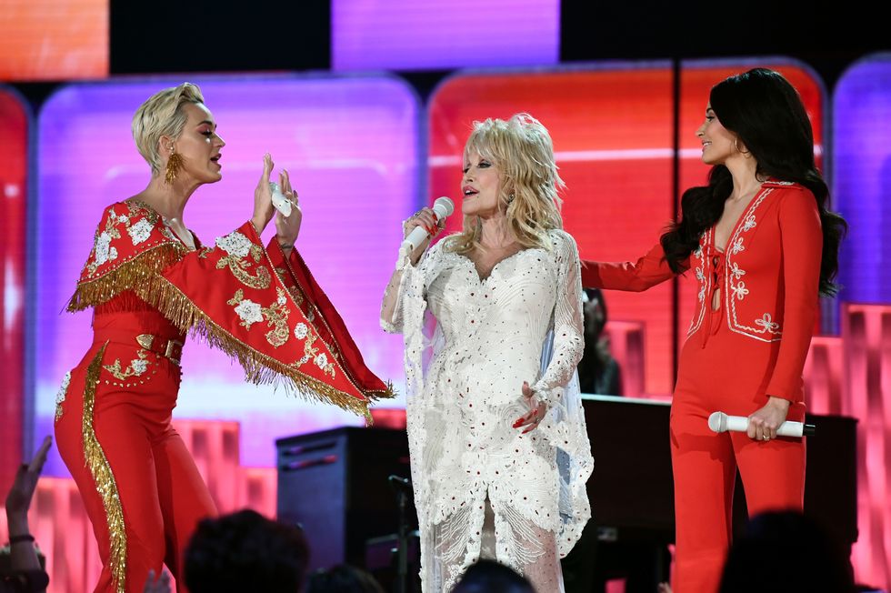 katy perry, dolly parton, and kacey musgraves stand on a stage each holding a white microphone, perry and musgraves wear red outfits with parton wearing a white dress