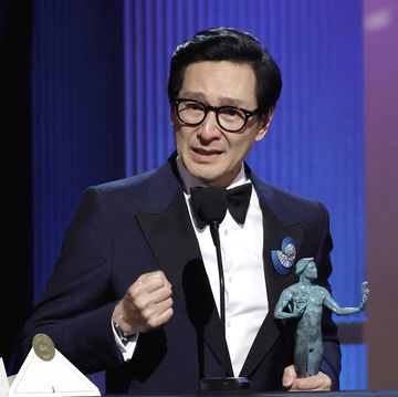 ke huy quan, wearing a black tuxedo, stands in front of a microphone on a stage, holding a sag award trophy in his left hand and clinching his right fist