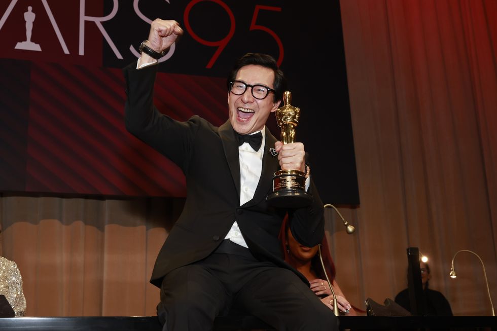 ke huy quan, wearing a black tuxedo, holds an academy award and raises his right fist into the air in celebration, sitting on a table in front of a banner that says oscars 95