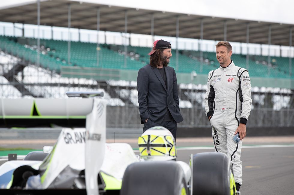 Keanu Reeves und Jenson Button reden miteinander und lächeln, während sie hinter einem bulligen Formel-1-Wagen auf einer Rennstrecke stehen