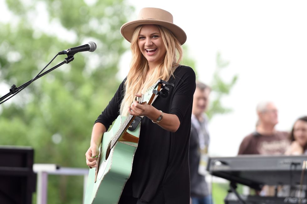 kelsea ballerini plays guitar and smiles at the camera while on a stage, she wears a tan hat and black dress, a microphone on a stand is on the left