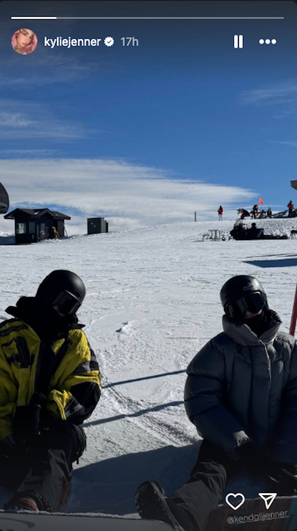 two individuals sitting on snow in winter clothing surrounded by a snowy landscape with ski lifts and other people in the background