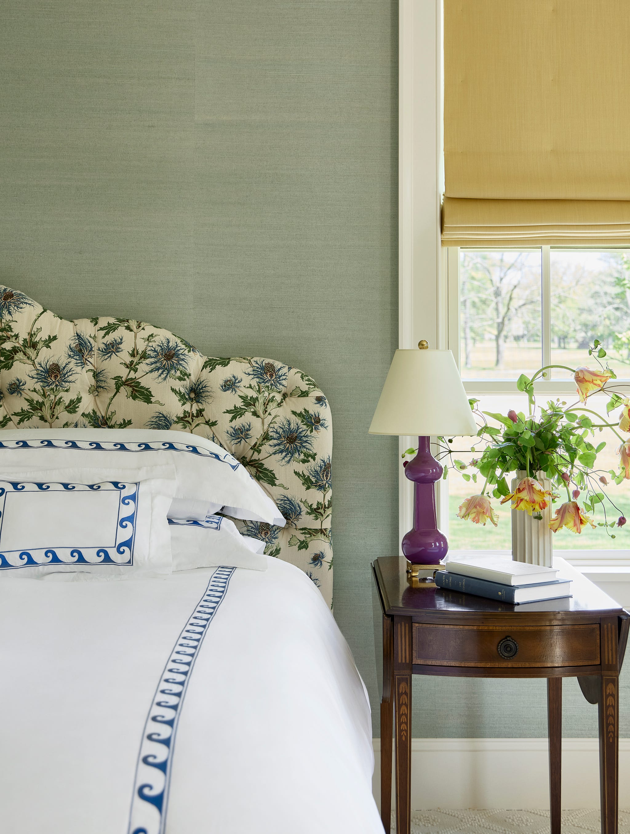 bed with white bedding with blue accents and floral headboard and an antique table next to it