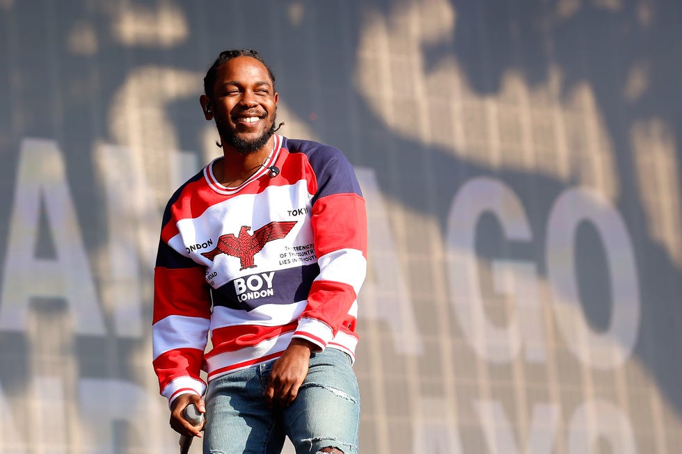 kendrick lamar smiles while standing outside on a stage, he holds a wireless microphone in one hand at his side, he wears jeans and a red, white, and blue striped shirt
