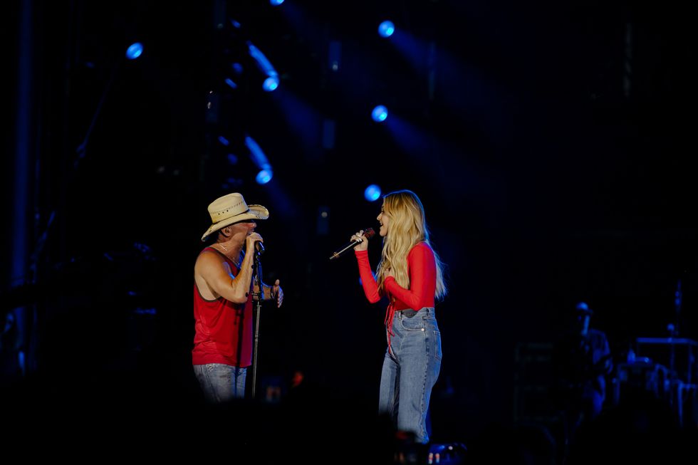 kenny chesney and kelsea ballerini stand on a stage and look at each other as they sing into microphones, both wear red shirts and blue jeans, chesney also wears a hat