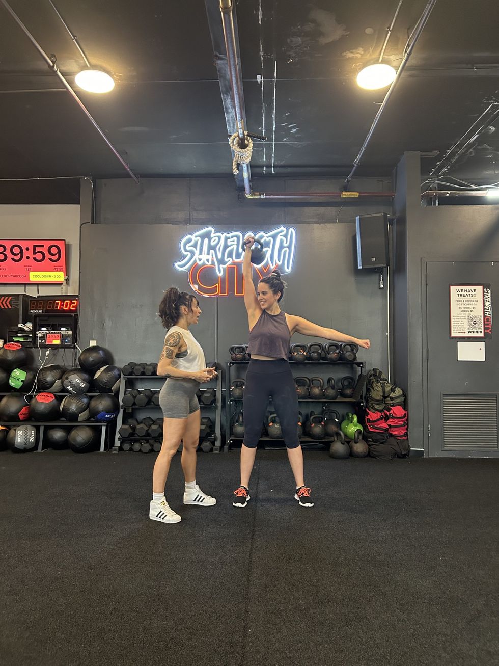 jordyn holds a kettlebell in the air while her trainer, alex, gives her pointers