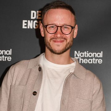 kevin clifton, a man stands looking at the camera and smiles, he has dark brown hair and beard, he wears glasses, a white tshirt with brown overshirt and trousers