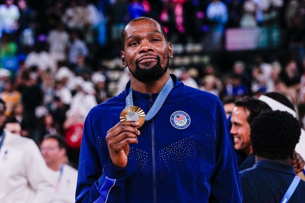 a man in a blue zip up jacket smiles while holding out a gold medal that hangs from his neck