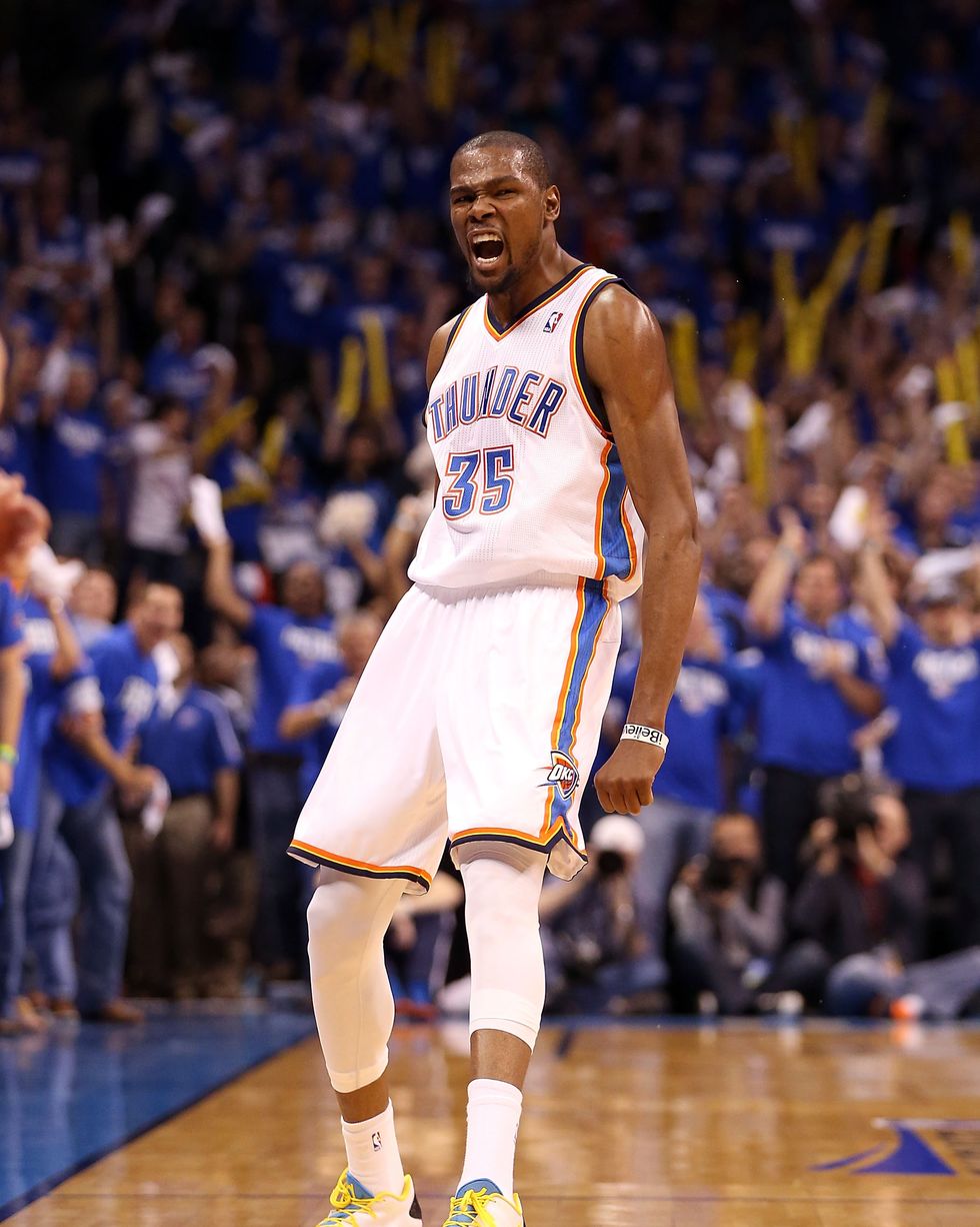 a man in a white baskbetball uniform with blue and orange details roars in delight as he stands on a court, fans are behind and to the left