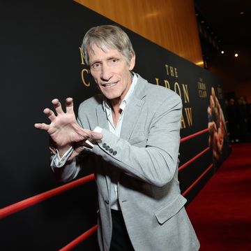 kevin von erich mimics the iron claw grapple for a photo while standing on a red carpet, he wears a gray suit jacket, white collared shirt and black pants, behind him is a large movie poster for the iron claw