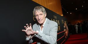 kevin von erich mimics the iron claw grapple for a photo while standing on a red carpet, he wears a gray suit jacket, white collared shirt and black pants, behind him is a large movie poster for the iron claw