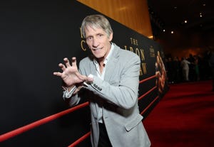 kevin von erich mimics the iron claw grapple for a photo while standing on a red carpet, he wears a gray suit jacket, white collared shirt and black pants, behind him is a large movie poster for the iron claw