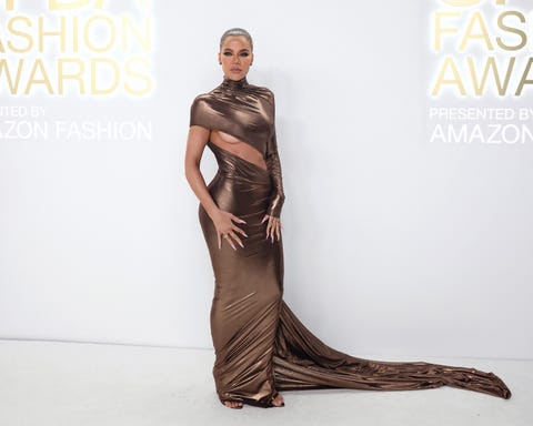 khloe kardashian, wearing a gold dress, standing in front of a white wall, looking directly at the camera