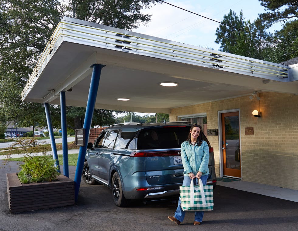 person holding a large checkered bag next to a parked kia carnival car and an entrance to a building