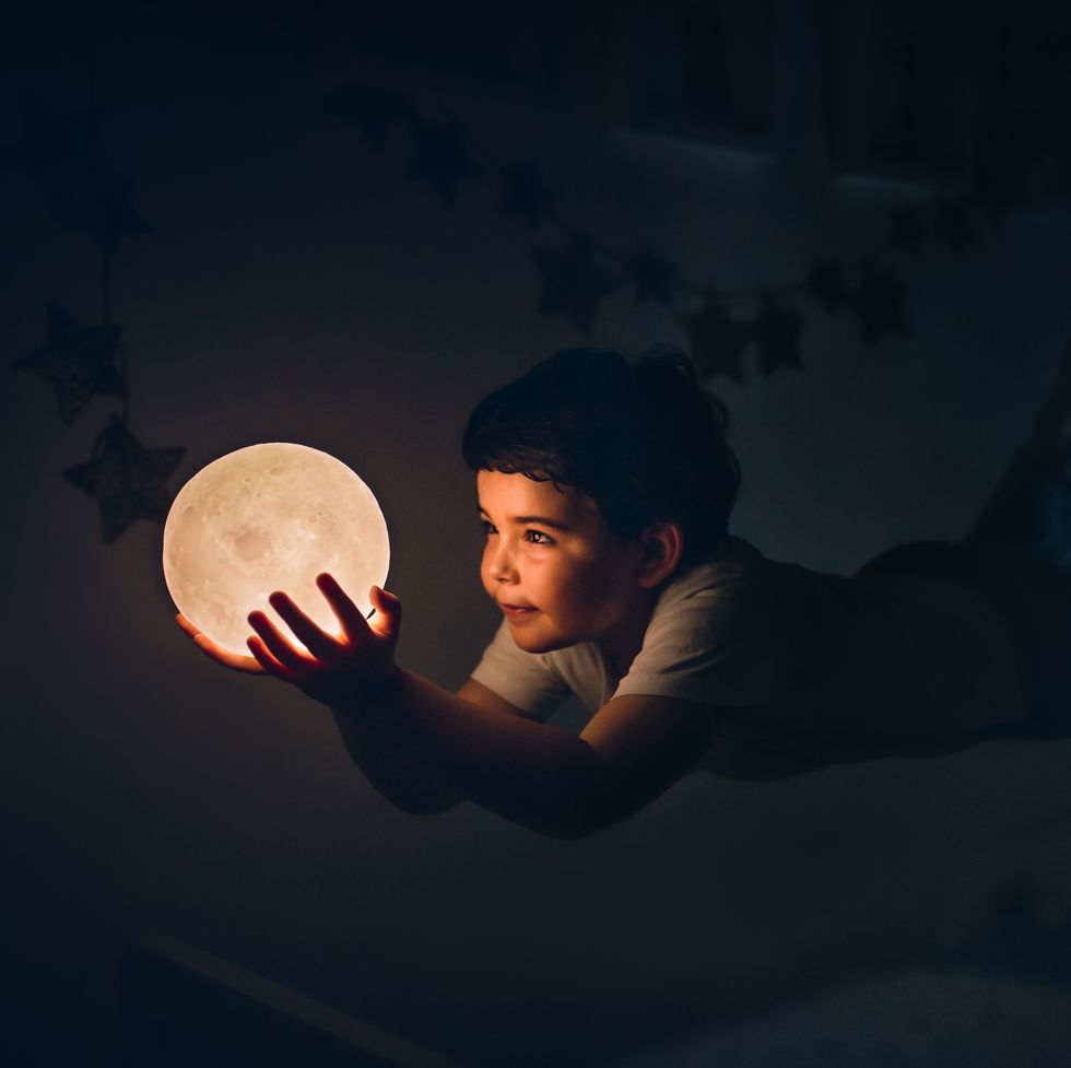 kid flying holding a moon lamp in the dark indoors