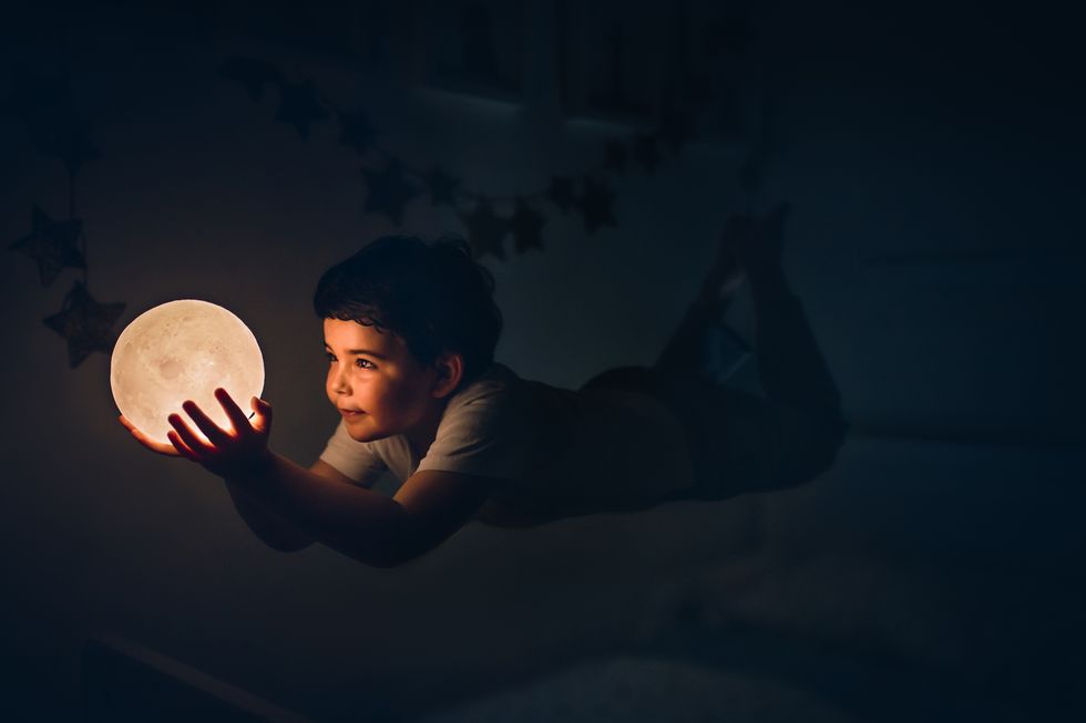 kid flying holding a moon lamp in the dark indoors