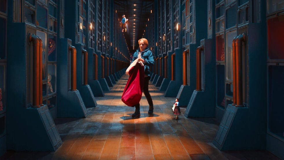 a kid stands in a long hallway of doors holding a red sack with a toy at his feet in a scene rom the claus family