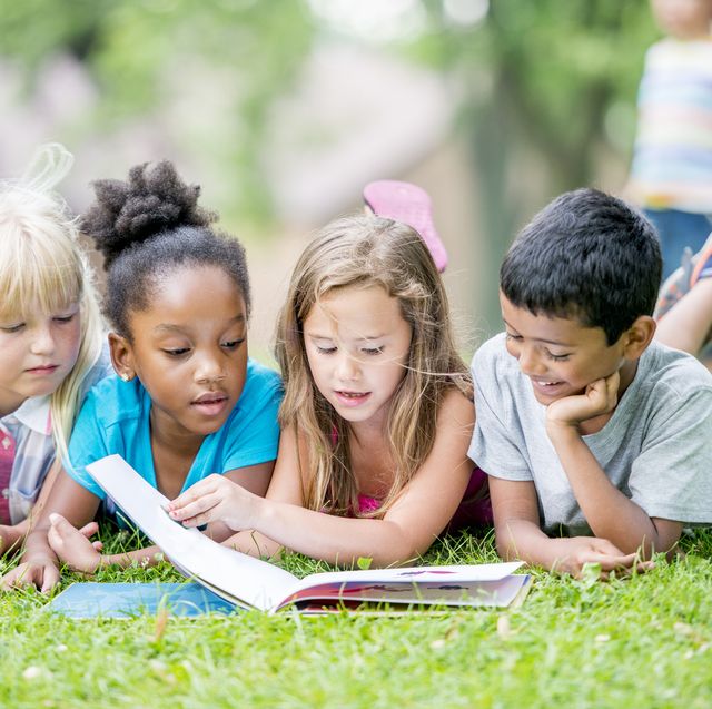 four kids laying on their bellies in the grass and reading a book together