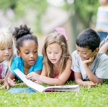 three young girls and a boy are outdoors during summer they are laying in the grass and reading a book together