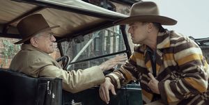 robert de niro and leonardo dicaprio in a scene from killers of the flower moon, de niro sits in an old car and reaches out to dicaprio who wears a brown, orange, and cream striped jacket, both men are wearing brown wide brimmed hats
