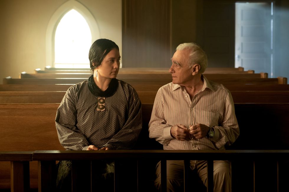 lily gladstone and martin scorsese in character for killers of the flower moon, they sit in a pew in an empty church and look at each other, she wears a gray long sleeve shirt, he wears a striped collared shirt