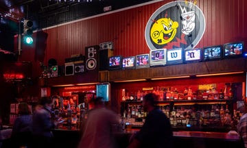 a vibrant bar scene featuring bar with various bottles displayed on shelves led lights provide ambient illumination creating a lively atmosphere on the wall, a colorful mural displays a cartoonish character with a drink vintage televisions with a retro design are mounted on the bar showcasing colorful visuals people can be seen enjoying the space contributing to the energetic vibe