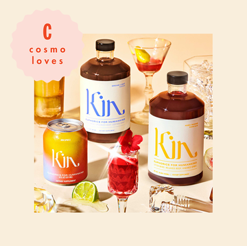 tablescape of non alcoholic cocktails made from kin euphorics, shown in glasses, bottles and cans on a peach background with a variety of fruit with a cosmo loves logo